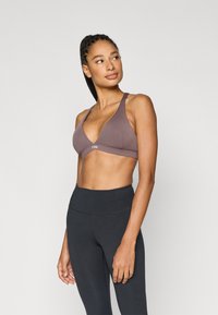 Σουτιέν σε απαλό καφέ χρώμα με βαθιά V-λαιμόκοψη, φαρδιές τιράντες και διακριτικό λογότυπο. Συνδυάζεται με μαύρο κολάν ψηλόμεσο για μια αρμονική εμφάνιση.