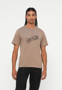 Maglietta in cotone marrone con maniche corte, caratterizzata da un design grafico nero. La vestibilità è rilassata e il collo è rotondo.