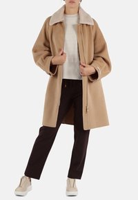 Cappotto in misto lana beige con colletto morbido e chiaro, vestibilità rilassata e due tasche frontali, indossato sopra un maglione chiaro e pantaloni scuri.