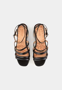 Sandales noires à lanières avec des semelles intérieures marron ornées de motifs de feuilles et des sangles à boucle autour de la cheville, vues de dessus sur un fond blanc.