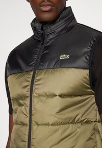Crni i maslinasto zeleni podstavljeni prsluk s visokim ovratnikom, punim zatvaračem i Lacoste logotipom na lijevoj strani prsnog koša. Sjajna tekstura s kvadriranim uzorkom.