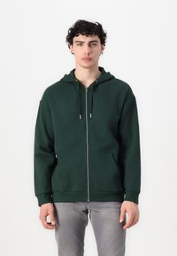 Hoodie zippé vert foncé avec une poche avant, capuche à cordon et poignets côtelés. Fabriqué à partir d'un tissu doux et texturé.