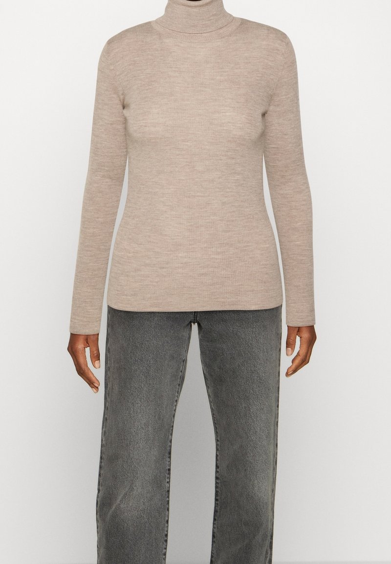 Pull à col roulé côtelé beige porté avec un jean noir délavé taille haute et jambes droites sur une personne à la peau foncée.