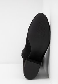 Botte en daim noir à bout pointu et semelle texturée, positionnée à la verticale contre des murs blancs qui se croisent.