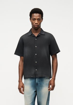 Άνδρας που φοράει μαύρο πουκάμισο με κοντό μανίκι και ανοιχτόχρωμο ξεθωριασμένο τζιν, στέκεται μπροστά σε λευκό απλό φόντο.