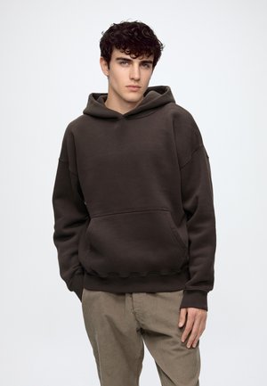 Junger Mann mit lockigem dunklem Haar, der einen dunkelbraunen Kapuzenpullover und beige Cordhosen vor einem schlichten hellen Hintergrund trägt.