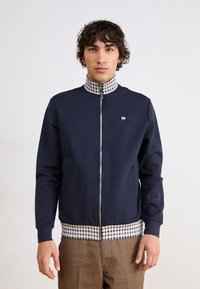 Navy zip-up jacket with a checkered collar and waistband. Features side pockets and a small logo on the chest. Smooth fabric texture.