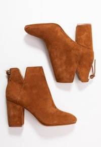 Ruske nubuk ankel støvler med blokkehæle, spidse tå, baglynlås og V-skarp detaljer på siderne, præsenteret på en hvid baggrund.