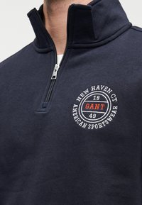 Ζακέτα σε ναυτικό μπλε χρώμα με φερμουάρ, φτιαγμένη από μαλακό ύφασμα. Διαθέτει κοντό γιακά και στρογγυλό λευκό κεντητό λογότυπο με κόκκινες λεπτομέρειες στο στήθος.