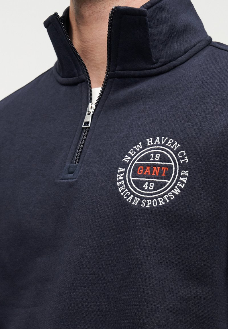 Felpa blu navy con zip realizzata in tessuto morbido. Presenta un colletto alto e un logo circolare ricamato bianco con dettagli rossi sul petto.