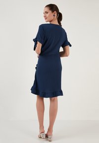 Sininen mekko, jossa on teksturoitu kangas, lyhyet hihat, joissa on nauhat, ja röyhelöhelma. Läpinäkyvät korot täydentävät asun.