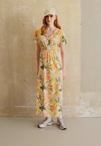 Virágmintás maxi ruha könnyű anyagból, narancs és zöld levélmintákkal. Bézs sapkával és ezüst sportcipővel kombinálva.