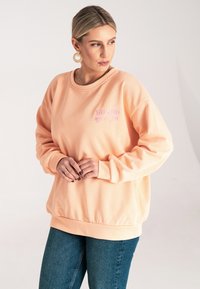 Brzoskwiniowy sweter w oversize'owym kroju, z prążkowanymi mankietami i dołem. Zawiera wyhaftowany różowy napis na lewej piersi. W połączeniu z dżinsami.