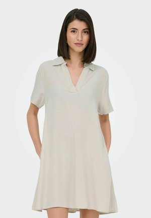 Ženska s temnimi lasmi, oblečena v ohlapno, svetlo bež kratko obleko s podložnim ovratnikom, stoji z rokami v žepih pred enobarvnim ozadjem.