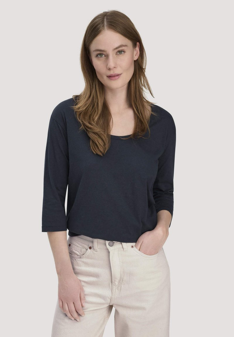Junge Frau mit langen hellbraunen Haaren, die ein schwarzes Langarmshirt und hellbeige Hosen trägt, stehend mit einer Hand in der Tasche.