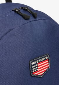 Mochila de marina con tejido texturizado, tiradores de cremallera negros y un parche que presenta el logo "SUPERDRY" junto a un diseño de bandera americana.