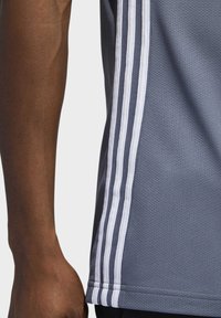 Šedé sportovní tričko s mřížkovou texturou, které má tři vertikální bílé pruhy po straně a zaoblený lem.