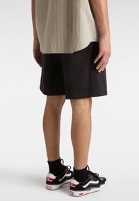 Pantaloni corti neri abbinati a sneaker Vans neri e rossi, con un calzino nero. La maglietta è chiara con righe verticali.