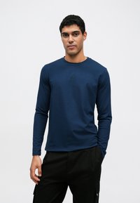 Μπλε μακρυμάνικη μπλούζα από μαλακό ύφασμα, με στρογγυλή λαιμόκοψη και μικρή λεπτομέρεια λογότυπου στο στήθος. Συνδυάζεται με σκοτεινό παντελόνι.