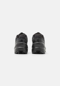 Une paire de chaussures de trail running noires vues de l'arrière, avec des semelles épaisses et des cols rembourrés sur un fond blanc uni.