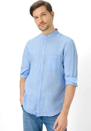 Mann mit hellbraunen Haaren, der ein hellblaues, langärmliges Hemd mit aufgekrempelten Ärmeln und blaue Jeans trägt, steht vor einem weißen Hintergrund.