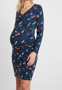 Gravidklänning i marinblått med ett livligt abstrakt mönster i röd, vit och svart. Tillverkad av mjukt tyg; har v-ringad halsringning och långa ärmar.