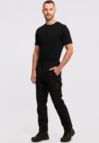 Schwarze, dehnbare Hosen mit Cargo-Taschen, einem schmalen Schnitt und einem blauen Logodetail. Getragen mit einem enganliegenden schwarzen T-Shirt und schwarzen Stiefeln.