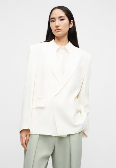 Λευκό διπλόμπουτο σακάκι με γιακά notch, υφασμάτινη υφή και μπροστινές τσέπες, συνδυασμένο με ανοιχτό πουκάμισο και ανοιχτό πράσινο παντελόνι.