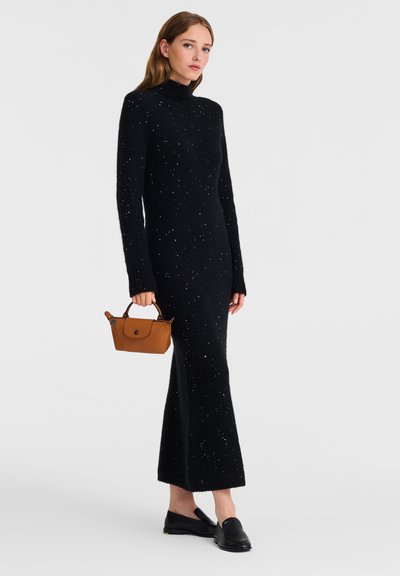 Schwarzes, langärmeliges Kleid mit Stehkragen und verstreuten weißen Sprenkeln, kombiniert mit einer braunen Handtasche und schwarzen Schuhen. Eng anliegende Passform, strukturierter Stoff.