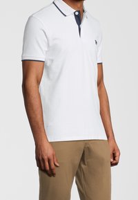 Vit pikétröja med marinblå krage, kort ärm och kontrasterande detaljer. Har en liten logotyp på bröstet. Slät bomullstyg.