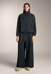 Temno siva trenirka z navpičnimi tankimi črtami. Zadrga na visokem ovratniku, elastični pas in široke hlače. Obele platformne čevlje v bež barvi.