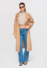 Femme portant un trench-coat beige, un crop top à carreaux, un jean bleu taille haute, une ceinture beige, des bottes en daim et des lunettes de soleil rondes, posant avec une main sur la hanche.