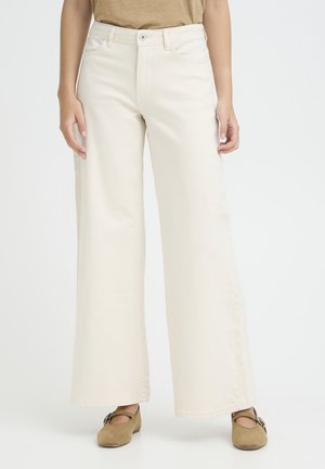 Persona che indossa pantaloni a vita alta, a gamba larga, color bianco sporco e scarpe Mary Jane marroni, con le mani rilassate ai lati, su uno sfondo semplice.