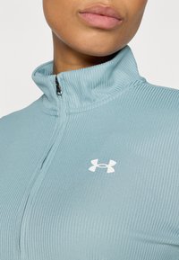 Jasnoniebieska, prążkowana sportowa kurtka zapinana na zamek z wysokim kołnierzem oraz białym logo Under Armour na klatce piersiowej. Zbliżenie na teksturę materiału.