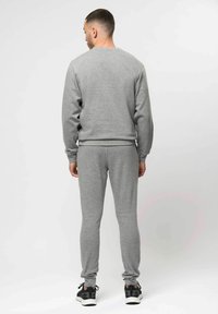 Man die wegkijkt, gekleed in een grijze sweatshirt en joggingbroek met zwart-witte sneakers, staand tegen een effen witte achtergrond.