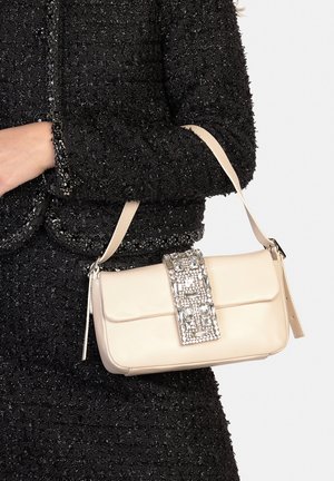 Bolso de mano de cuero beige con un cierre acentuado en plata y adornos de gemas, llevado sobre una chaqueta negra texturizada.