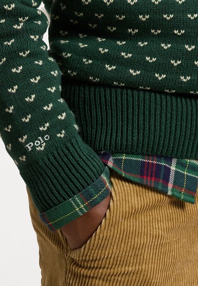 Sweater verde tricotado com padrões brancos em forma de coração, punhos canelados e o logótipo "Polo". Por baixo, uma camisa quadriculada em tons de azul, verde e vermelho.