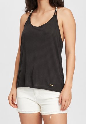 Femme portant un haut noir sans manches à fines bretelles et un short en jean blanc effiloché, debout devant un fond uni.