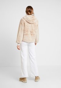 Beżowa kurtka z sztucznego futra z kapturem, o luźnym kroju, miękkiej teksturze i długich rękawach. W połączeniu z białymi spodniami i masywnym, brązowym obuwiem.