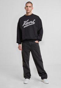 Sort sweatshirt med hvid skriftet logo, afslappet pasform, ribbede manchetter, parret med løstsiddende mørkegrå bukser og hvide sneakers.