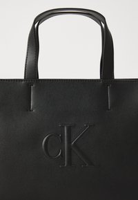 Borsa a tracolla nera in pelle con logo "cK" in rilievo, manici resistenti e texture liscia. Presenta un design minimalista e dettagli di cucitura discreti.
