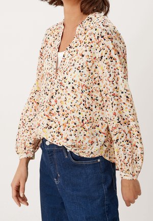 Žena v volné dlouhém rukávu halenky s abstraktním vícebarevným potiskem, oblečená k tmavě modrým džínům, stojící před jednobarevným pozadím.