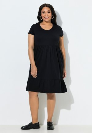 Mujer con vestido negro de manga corta hasta la rodilla, con cintura y dobladillo fruncidos, mocasines negros y grandes pendientes de aro, sonriendo a la cámara.