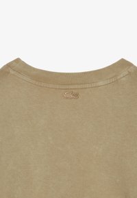 Camiseta de algodón beige con cuello redondo; presenta un escote texturizado y un pequeño logotipo bordado en la parte posterior.