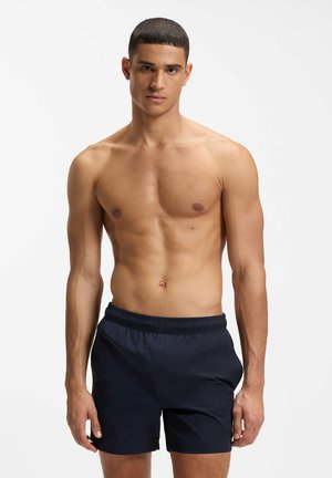 Jonge man met kort haar, gekleed in marineblauwe zwemshorts, tegen een effen witte achtergrond.