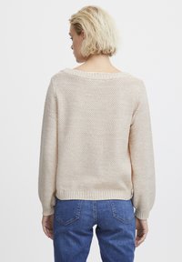 Beiger Strickpullover mit strukturiertem Muster, weitem gerippten Ausschnitt und leicht gepufften Ärmeln, kombiniert mit blauen Jeans.