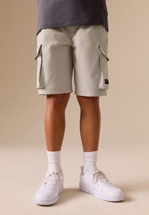 Persona que lleva pantalones cortos cargo beige, calcetines blancos y zapatillas blancas, de pie contra un fondo beige liso.