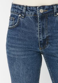 Pantalones vaqueros de mezclilla azul oscuro con un diseño ajustado, que presentan un estilo clásico de cinco bolsillos, costuras amarillas contrastantes y un cierre de botón de metal.