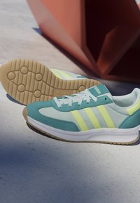 Hellblaue und gelbe Adidas-Sneaker mit weißen Schnürsenkeln und Gummisohlen, die auf einem glatten Betonboden in der Nähe einer roten geometrischen Struktur liegen.