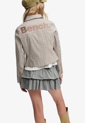 Femme portant une veste beige à carreaux avec le logo "Bench.", jupe grise à volants, casquette vert olive et bottes couleur beige, vue de dos.